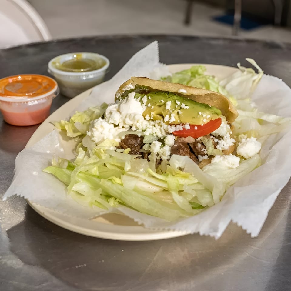 Arepa with meat, avocado, tomato, and cheese, served with lettuce and two sauces on a metal table.