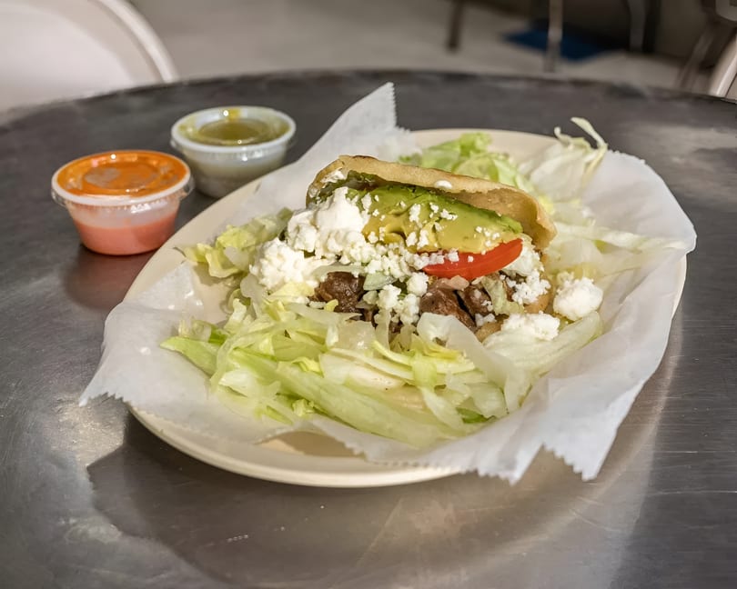 Arepa with meat, avocado, tomato, and cheese, served with lettuce and two sauces on a metal table.