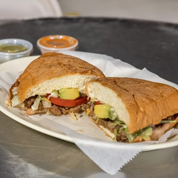 A torta sandwich, cut in half, with meat, lettuce, tomato, and avocado, served with two sauces.