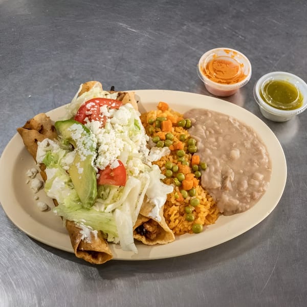 A plate of taquitos topped with lettuce, tomato, avocado, and cheese, with rice and beans.