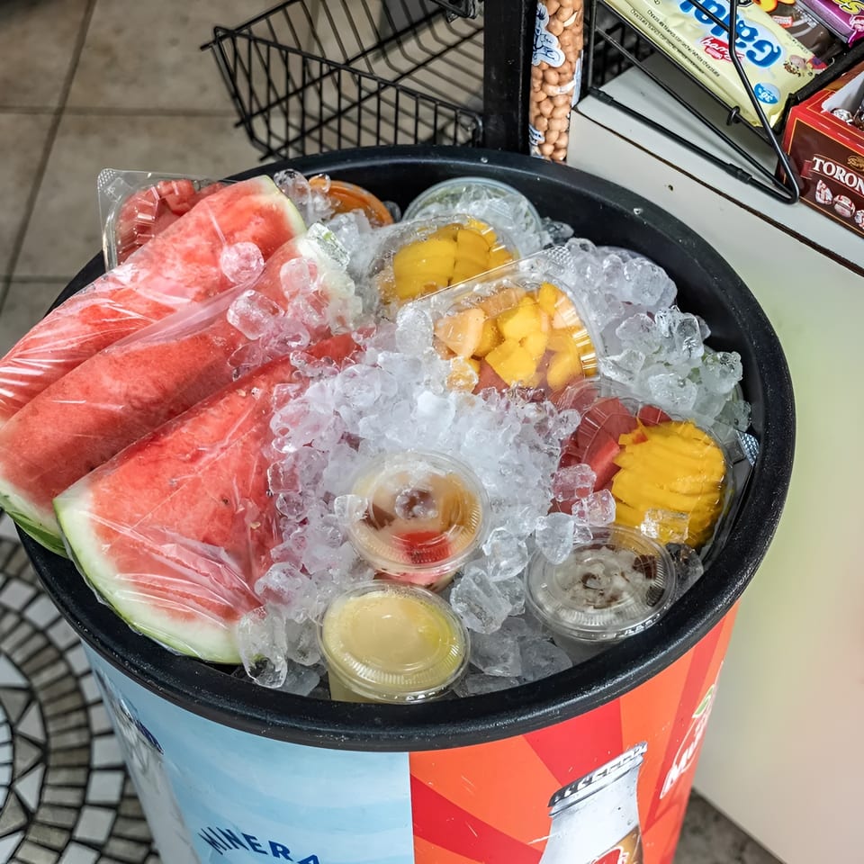 Watermelon slices and fruit cups on ice in a cooler, with snacks on a shelf nearby.