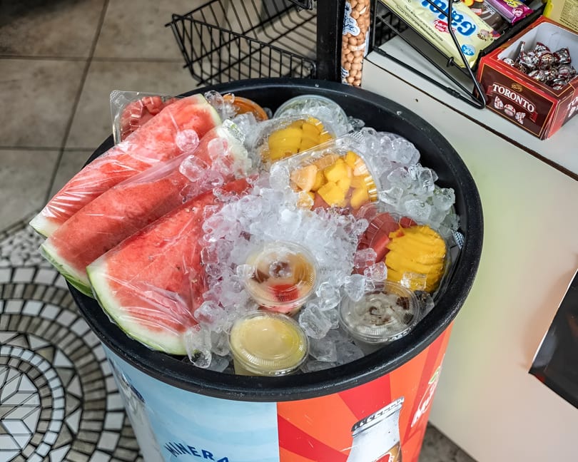 Watermelon slices and fruit cups on ice in a cooler, with snacks on a shelf nearby.