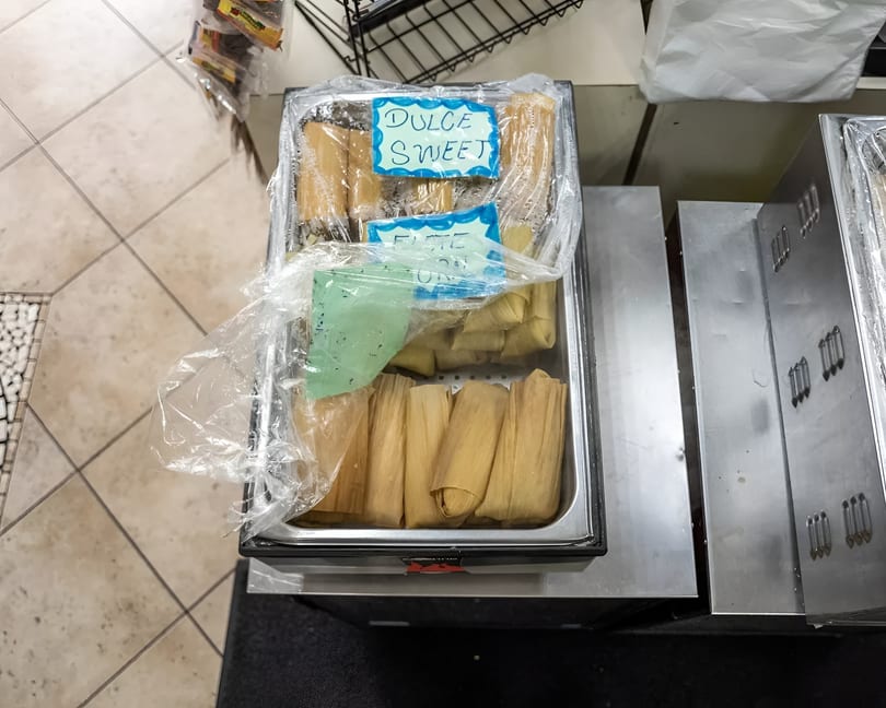 A metal tray filled with tamales, some labeled "Dulce Sweet," covered in plastic wrap.
