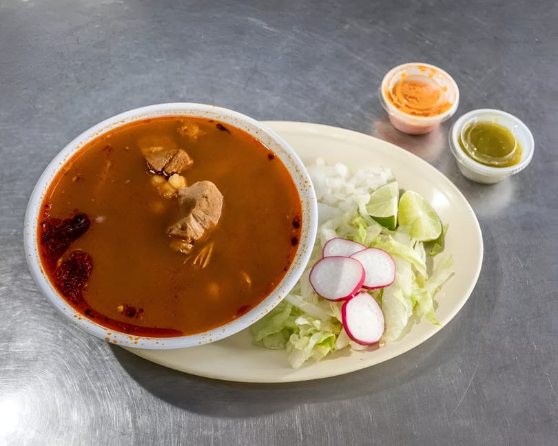 Hearty red pozole soup with pork and hominy, served with a side of lettuce, radishes, and lime.