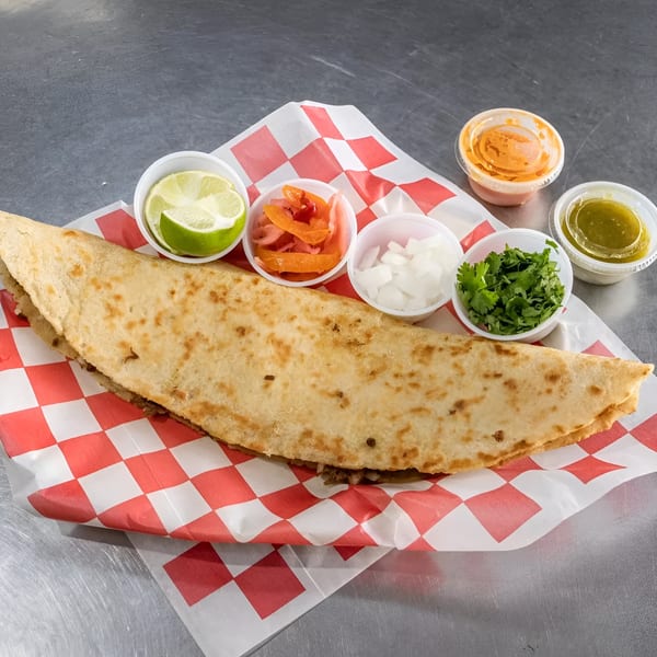 Large quesadilla on red and white checkered paper, with sides of lime, pickled onions, cilantro, and sauces.