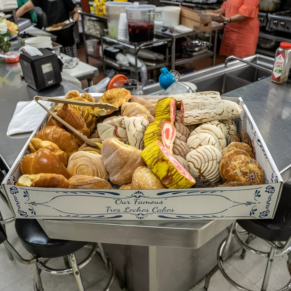 A large box filled with various Mexican pastries, including conchas and colorful cakes, on a counter.