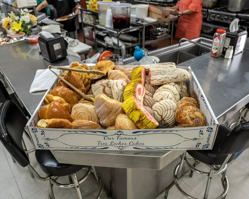 A large box filled with various Mexican pastries, including conchas and colorful cakes, on a counter.