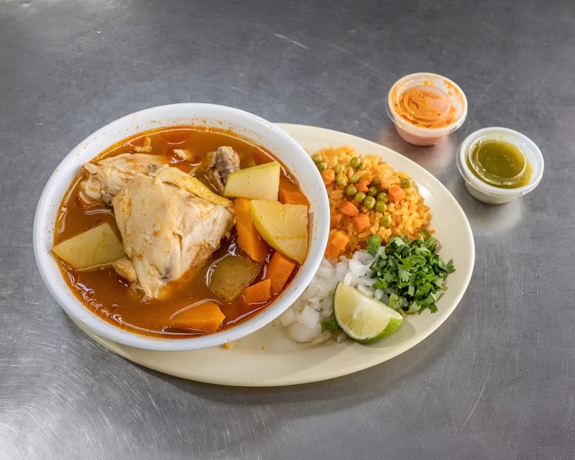 A bowl of chicken soup with vegetables, rice, and a side of chopped onions, cilantro, and lime.